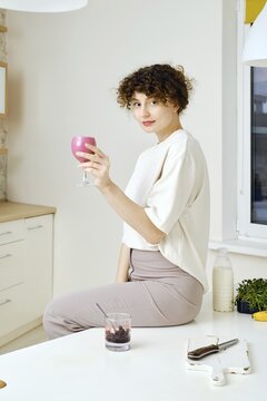 Cute woman sits on a kitchen table and holds a glass of black currant smoothie in hand