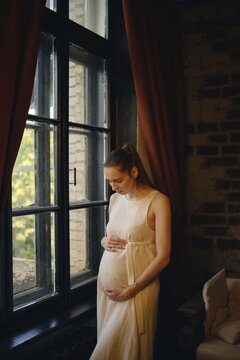 A pregnant woman stands by a large window in her home, gently cradling her belly and a listens to the movements of a baby in the womb
