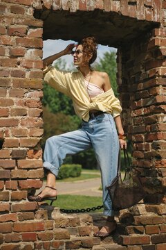 A model outfits herself in a trendy summer ensemble featuring a light yellow button-up shirt and high-waisted jeans while leaning against a rustic brick wall