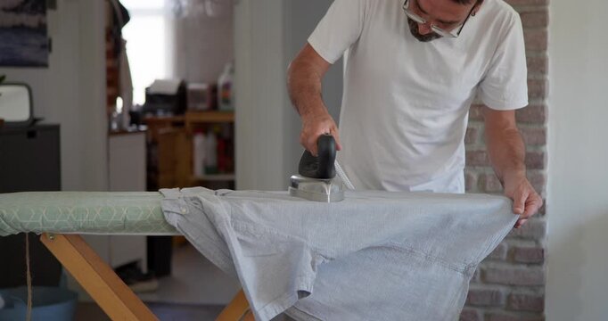 Man Ironing Clothes: A man wearing a white t-shirt and glasses is diligently ironing a grey t-shirt on an ironing board, engaged in a common household chore.