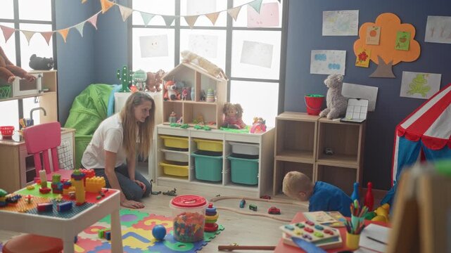 Woman kneels beside a little boy pushing toy train along wooden track and stacking colorful blocks on foam mat in building; nurturing play.