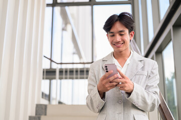 Fototapeta premium Smiling young nonbinary Thai business professional texting on smartphone on office stairs