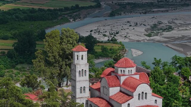Aerial view of a traditional Orthodox church with red tiled domes and a bell tower overlooking the Vjosa River and green valley in Tepelene, Gjirokaster County, Albania.