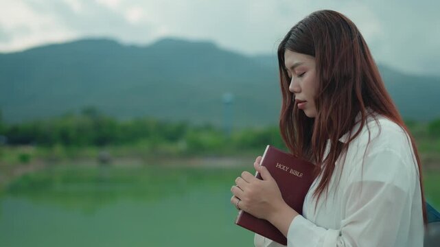 Faith concept: young Asian Christian woman embracing Holy Bible with eyes closed, praying outdoors by peaceful lake and mountain, symbolizing hope, peace and spirituality. 