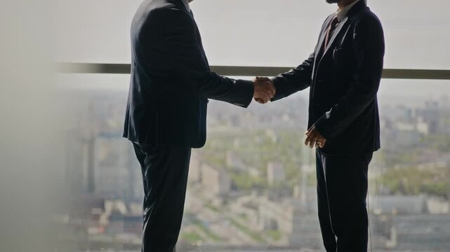 Tilt up shot of senior Caucasian businessman and young Indian partner standing by office window, shaking hands, and discussing partnership. Clip is ideal for corporate communication