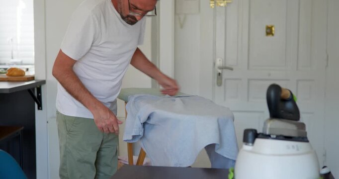 Man Ironing Shirt at Home: A man in casual attire is carefully ironing a light blue shirt, with an ironing board and iron visible, suggesting a domestic chore.