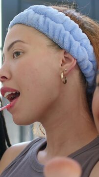 Vertical video: Applying woman in blue headband and gray top pressing lip wand at vanity prepping