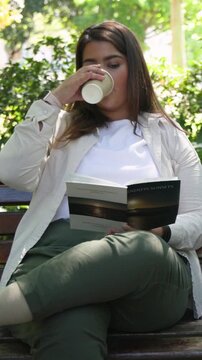 Vertical video: Reading woman in beige on park bench holding book and paper cup as walker crossing