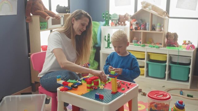 Woman and little boy placing colorful toy bricks on a small play table, woman places a brick with her hand in a building playroom filled with toys; bonding.