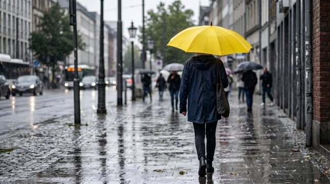 A woman in a navy raincoat walks with a yellow umbrella on a rainy city street, reflecting the grey mood and wet pavement.