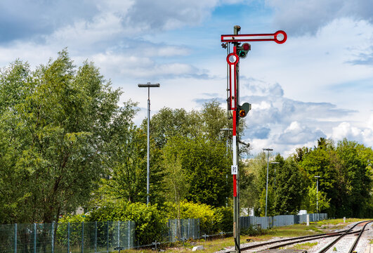 Historic railway semaphore signal with red and white arm next to tracks, traditional mechanical train traffic signaling system with green trees and cloudy sky background.