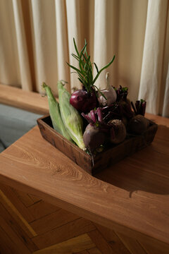 Fresh organic vegetables including corn, beetroot, and red onions with garlic in a rustic wooden tray on a table. Cozy kitchen interior