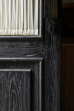 Close-up of an old vintage black wooden door with a weathered texture, a keyhole, and a small glass window with a white curtain. Antique interior details