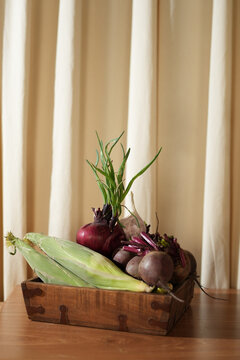 Fresh organic vegetables including corn, beetroot, and red onions with garlic in a rustic wooden tray on a table. Cozy kitchen interior