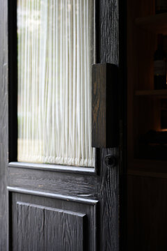 Close-up of an old vintage black wooden door with a weathered texture, a keyhole, and a small glass window with a white curtain. Antique interior details