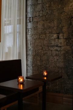 Burning small candles on tables in a cozy cafe with an old brick wall and white curtain. Moody elegant restaurant interior