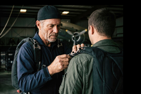 Senior military instructor checking parachute harness of a young pilot inside hangar facility