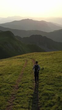 Young man with a backpack hiking on a scenic mountain path. Beautiful golden hour light illuminates the lush green hills and distant peaks