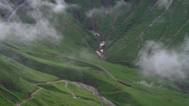 High Altitude Aerial Drone View of Cogwheel Train on Swiss Alpine Railway with Green Meadows and Clouds