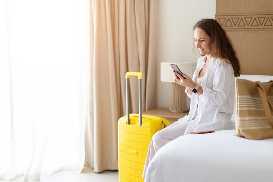 Smiling woman sitting on hotel bed using smartphone beside bright yellow suitcase, checking travel information in a modern bedroom during a leisure or business trip