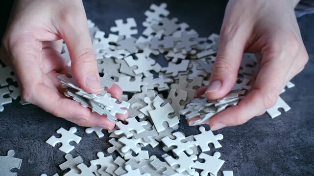 Close up view of hands mixing and sorting through a pile of white jigsaw puzzle pieces on a dark gray surface, searching for the right connection to solve the challenging mind game
