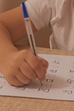 Close up horizontal shot of a child holding a pen and practicing writing english alphabet letters on a white paper sheet with dotted lines at a wooden desk