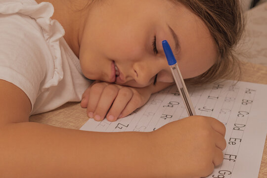 Horizontal close up view of an exhausted child sleeping soundly on a desk while holding a blue ballpoint pen over a white copybook during english alphabet practice