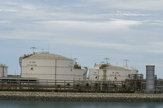 large white dome-roofed industrial storage tanks for propylene oxide chemical storage behind complex pipelines and a stone breakwater on a waterfront industrial terminal under a cloudy sky