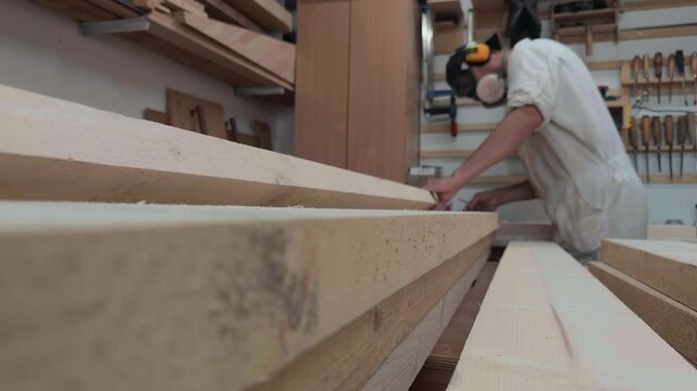 Cinematic rack focus shifting from professional woodworker in full PPE measuring wooden board to extreme macro detail of raw timber plank texture in bright rustic garage workshop authentic broll