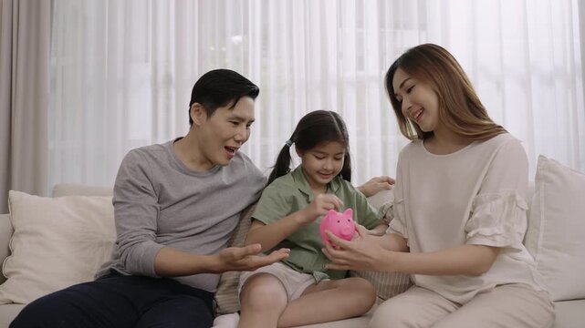 Happy Asian family saving money concept. Smiling father and mother with child girl sitting saving money into a pink piggy bank together for their daughter's future on sofa in the living room at home. 