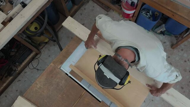 Anonymous professional artisan in full safety ppe gear ripping raw timber plank on heavy table saw machinery in bright fabrication workshop authentic high angle overhead top down b-roll shot