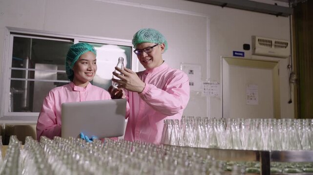 Two Asian quality control inspectors in uniform standing, using a laptop computer to analyze and inspect empty glass beverage bottles on moving conveyor belt in production line at industrial factory. 