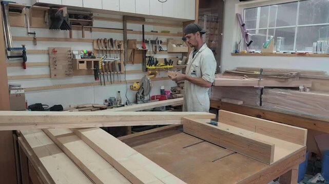 Focused joiner taking try square from tool rack measuring wooden block at workbench in bright garage studio authentic manual labor woodworking production process cinematic documentary wide shot