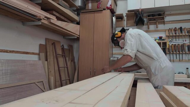 Focused joiner wearing white coveralls and cap measuring and inspecting raw timber planks at lumber stack in bright rustic workshop environment waist level low angle perspective authentic b-roll