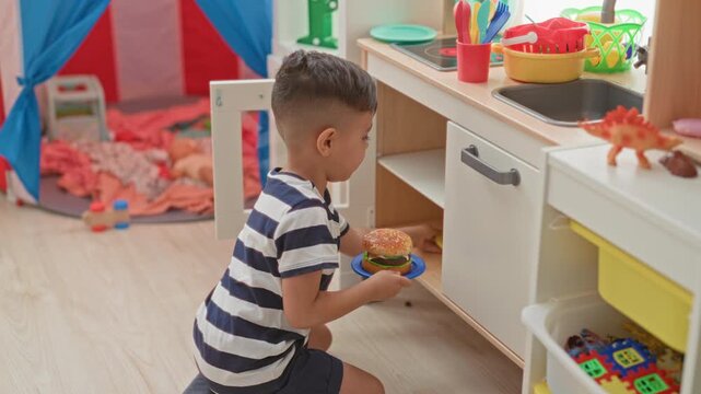 Little boy in striped shirt kneels and holds two toy hamburgers on plates at a play kitchen shelf inside a building; playful.