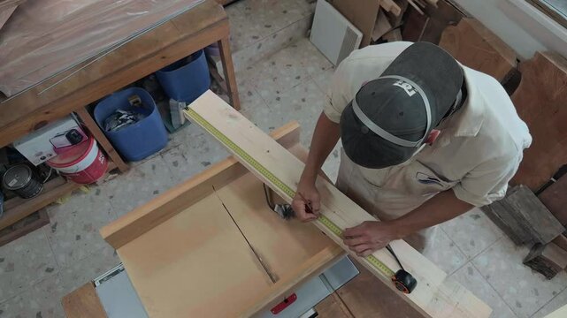 Focussed carpenter in white coveralls carefully measuring and marking raw timber plank with pencil and wooden folding ruler in rustic workshop handheld close up documentary style authentic broll