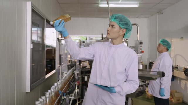 Asian quality control inspector in a uniform food and drink factory holding a tablet inspecting fruit juice bottles while moving on conveyor belt in beverage production line at manufacturing industry.