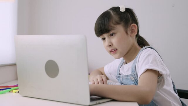 Cute Asian girl student sitting at desk using a laptop computer for studying online learning via video calling with remote teacher in a home school. Homeschooling education and new normal concept. 