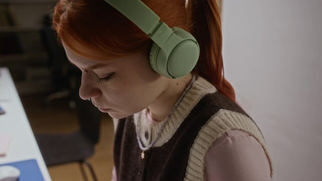 Dolly in shot of Caucasian teenage girl wearing headphones typing on keyboard while studying in library. Footage ideal for online education platforms, e-learning ads, and student lifestyle promos