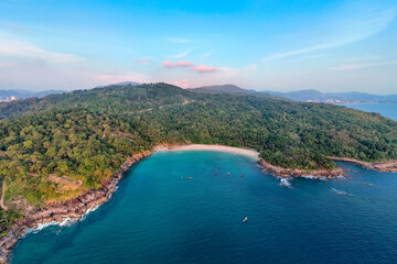 Obraz premium Golden hour light bathes Freedom Snorkel Beach Phuket Thailand as the sun sets over the ocean. Boats rest in the calm, turquoise bay, hugged by dense jungle foliage
