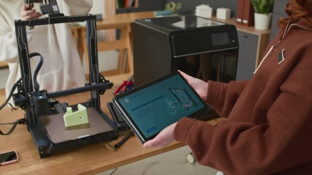 Unrecognizable girl holding digital tablet with instructions while working on 3D printer with her classmate during practical lesson in school