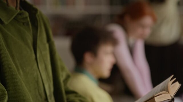 lose up shot of African American teenager with dreadlocks and glasses reading book in library with classmates, tilt up camera movement. Footage for educational marketing, student lifestyle campaigns