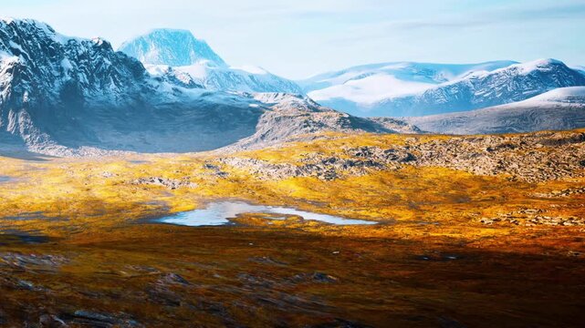 A vast mountain landscape with rugged terrain. A small lake is visible among yellow grass and rocky areas. Snow capped peaks rise in the background under a clear sky.