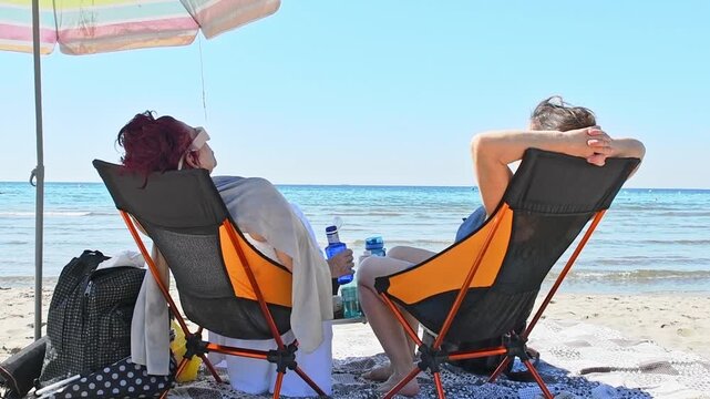 An elderly and young woman relax in sun loungers under an umbrella, enjoying the sea view on a summer vacation day. Accessories for a summer vacation. Tourism in a hot country