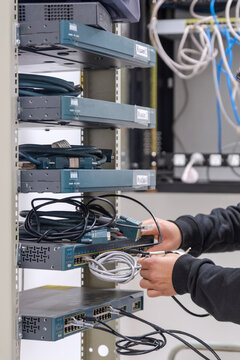 Close-up of a technician's hands plugging a blue ethernet network cable into a switch inside a rack with multiple network devices.