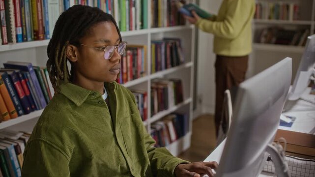 Medium shot of African American teenager with dreadlocks typing on computer in library with classmate standing near bookshelf. Clip ideal for educational marketing and school promotional videos