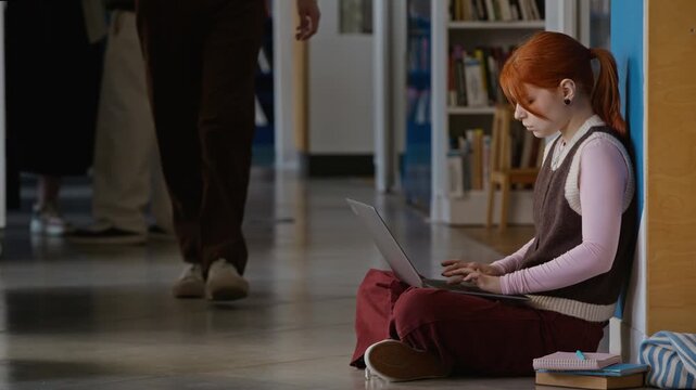 Full length shot of Caucasian teenage girl sitting on library floor and typing on laptop while students walk by in background. For educational marketing and online learning promotional campaigns