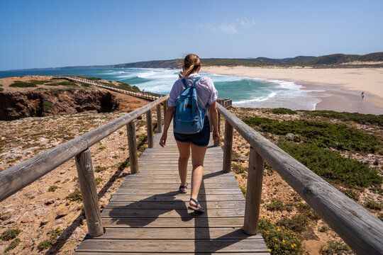 Visiting Algarve coast in Portugal. Beautiful middle-aged woman tourist walking down wooden stairs to sandy Bordeira beach and enjoying vacations over ocean on summer day. Back view