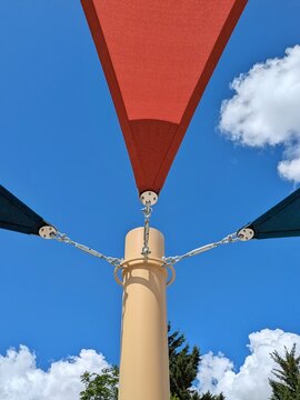 Vista de cerca de tensores con cables de metal en la parte superior de postes de sujeci&oacute;n de parasoles triangulares de exterior, de color rojo y azul, en plaza p&uacute;blica	