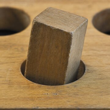 A square wooden peg attempts to fit into a round hole, showing worn edges.